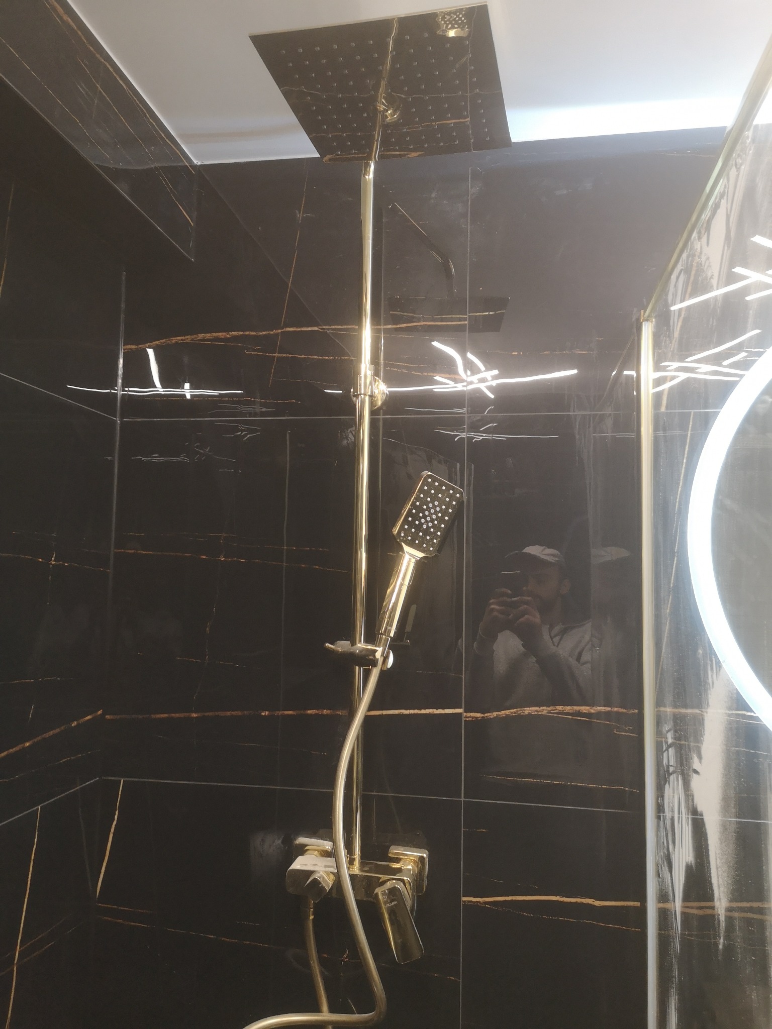Kabina prysznicowa z czarnymi płytkami imitującymi marmur ze złotymi żyłami, złota armatura, deszczownica sufitowa i oświetlenie LED w lustrze odbijające fotografa.