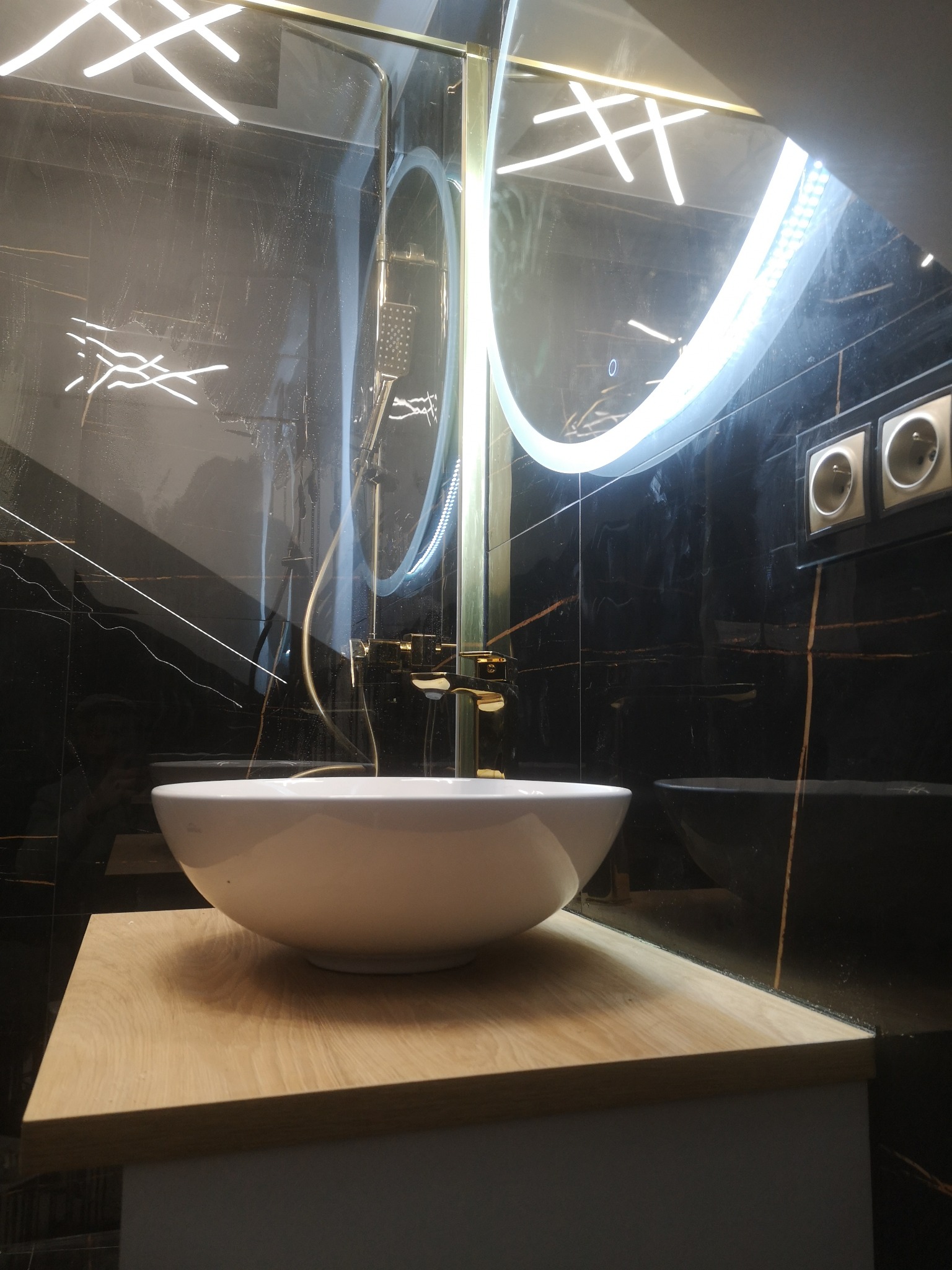 Nowoczesna łazienka z czarnymi płytkami imitującymi marmur, okrągłym lustrem z podświetleniem LED, białą umywalką nablatową na drewnianym blacie, złotą armaturą i szklanym panelem prysznicowym.