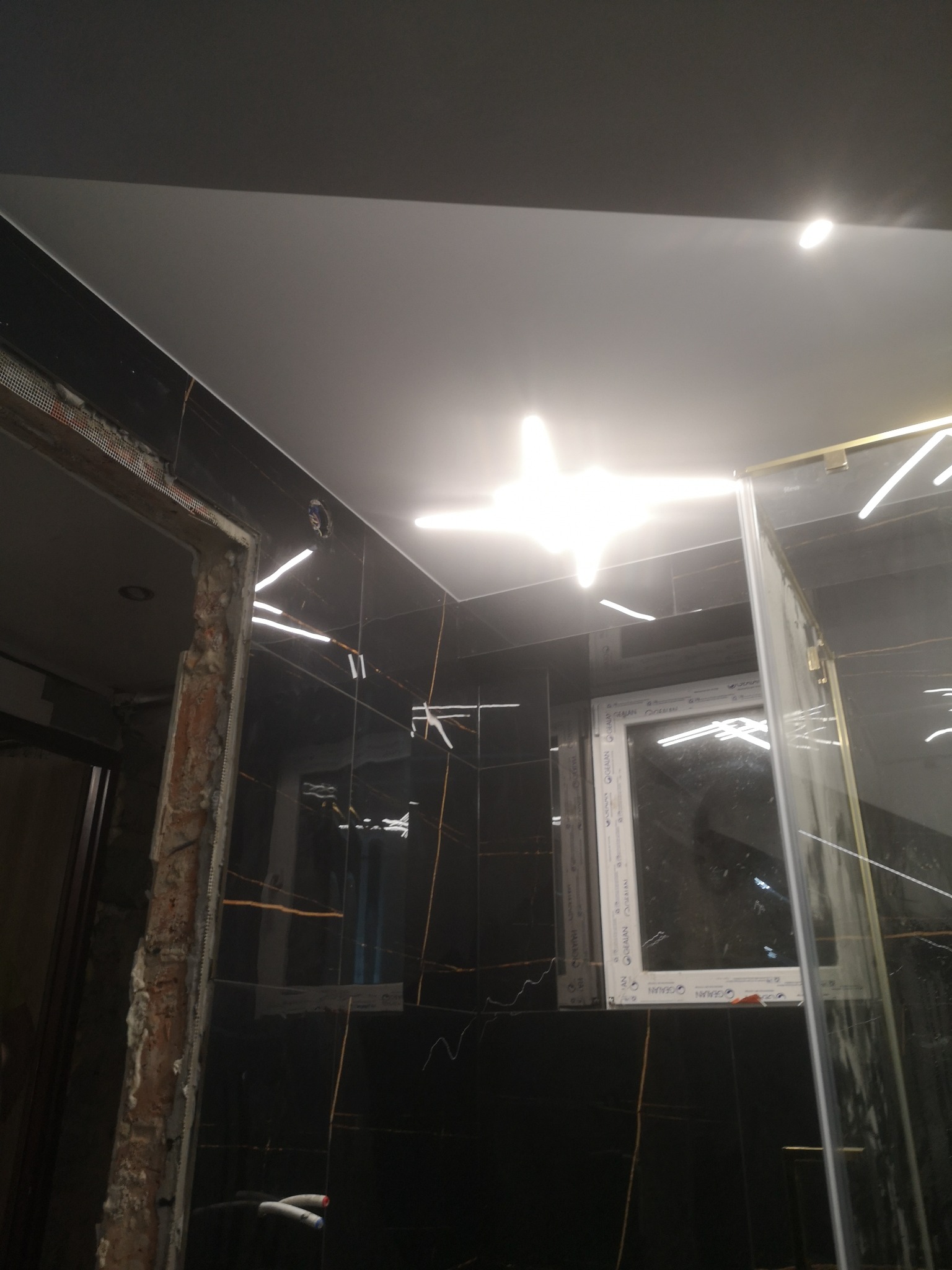 Częściowo wykończona łazienka z czarnymi płytkami z złotymi żyłkami, widoczne fragmenty instalacji elektrycznej i ramy drzwiowej w trakcie prac wykończeniowych, oraz kabina prysznicowa z elementami...