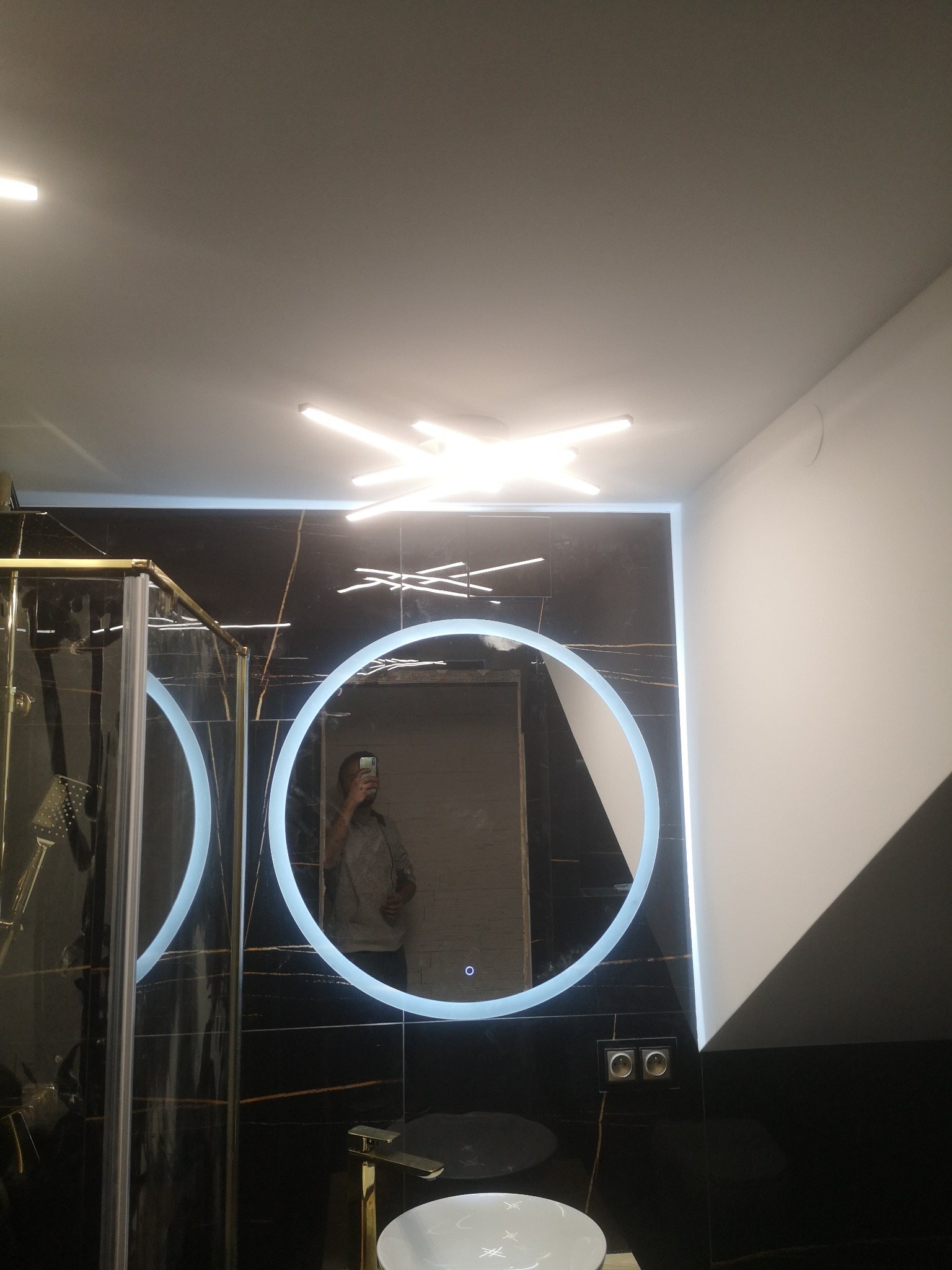 Nowoczesna łazienka z czarnymi płytkami imitującymi marmur, okrągłym lustrem z podświetleniem LED, złotą armaturą i kabiną prysznicową ze złotymi profilami, oświetlona lampą sufitową w kształcie...