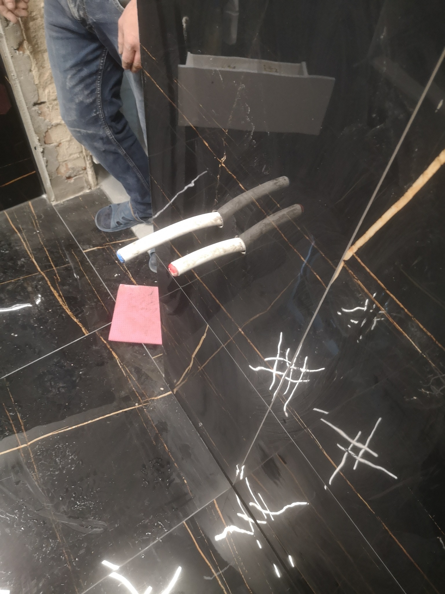 Czarna, błyszcząca płytka podłogowa z złotymi żyłkami, z widocznymi śladami po fugowaniu, różowa gąbka do czyszczenia i fragment instalacji hydraulicznej z rurami PCV