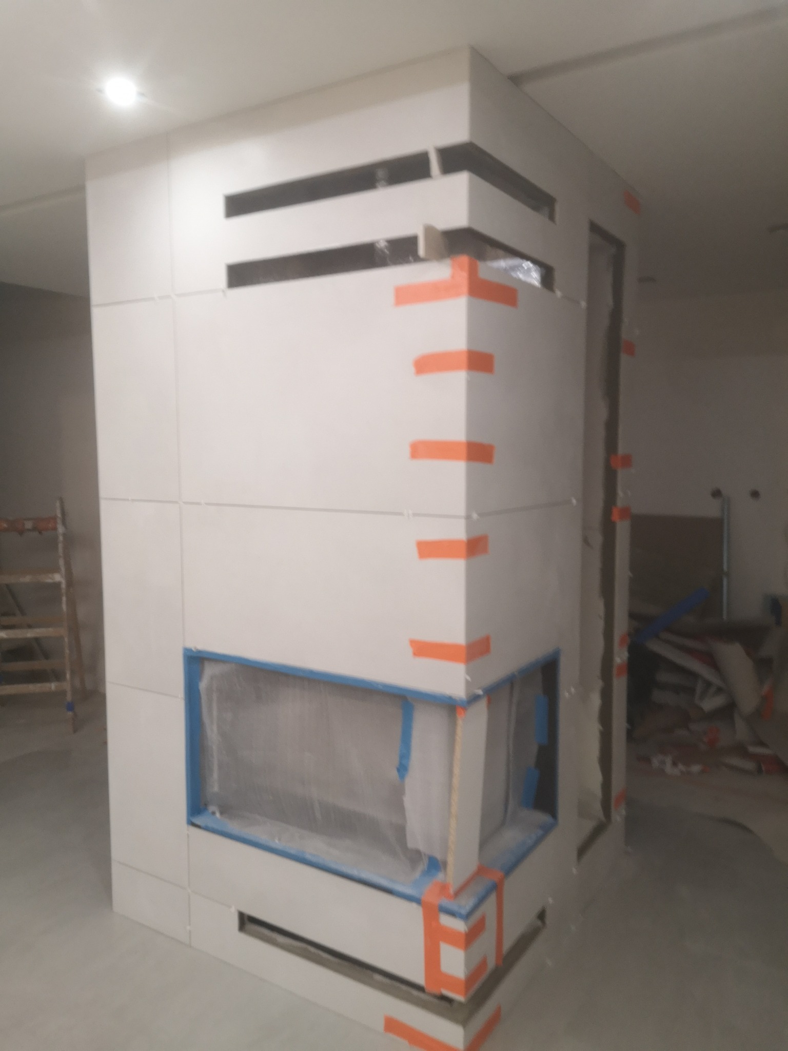 Kanciasta zabudowa z białych płytek, zabezpieczona pomarańczową taśmą malarską, w trakcie prac wykończeniowych, widoczne zabezpieczone folią otwory.