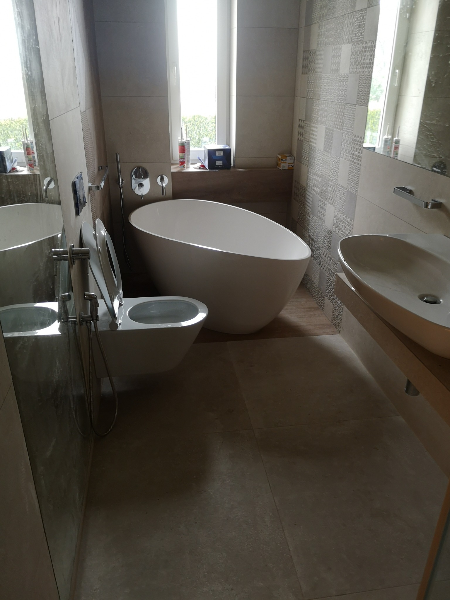 Nowoczesna łazienka z wolnostojącą wanną, wiszącą toaletą z bidetką, umywalką nablatową i mozaiką na ścianie, widoczne silikony na parapecie okna.