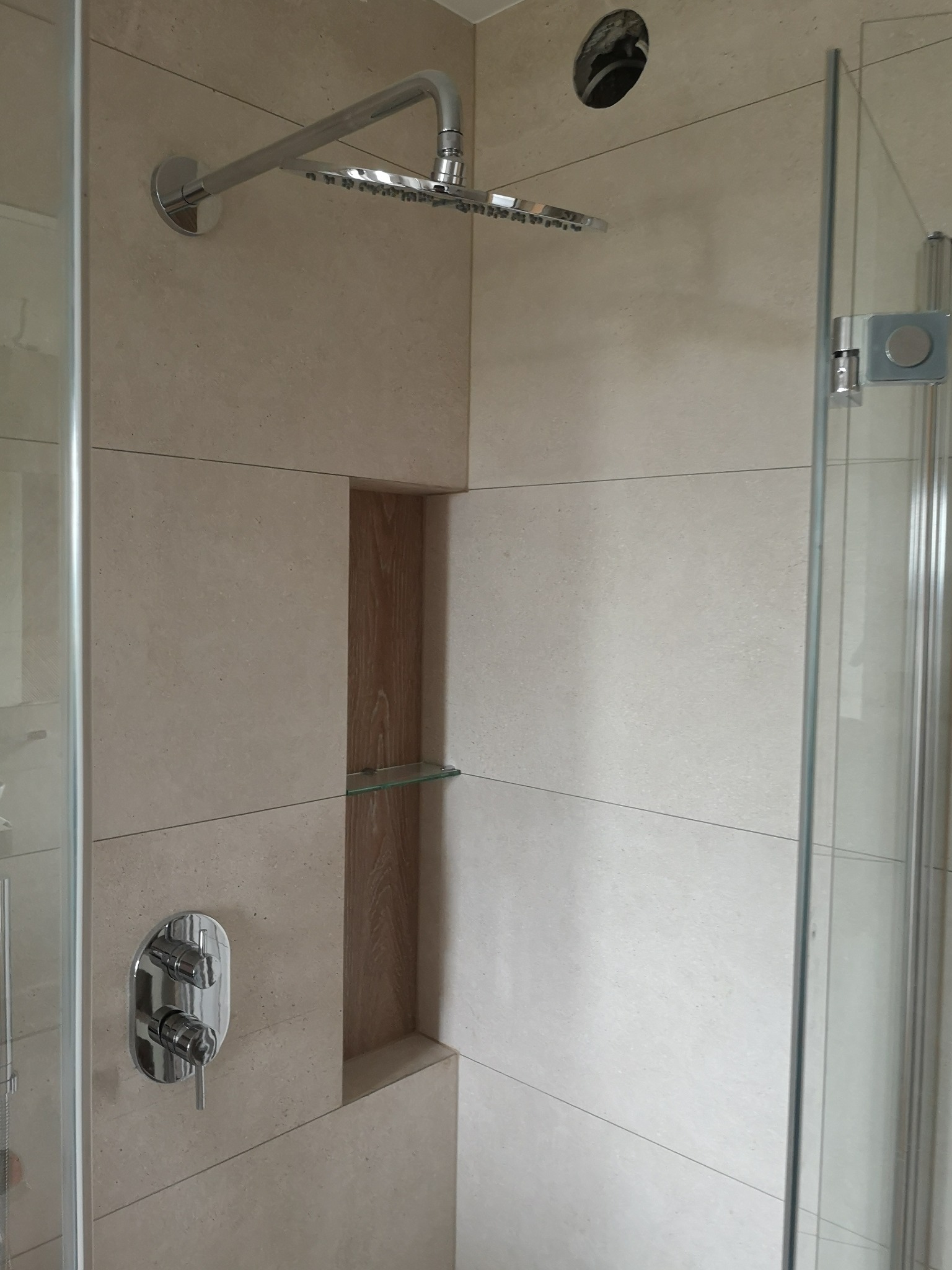 Beżowa kabina prysznicowa z deszczownicą, wbudowaną półką z drewnianym wykończeniem i szklaną półką, oraz chromowaną baterią podtynkową.