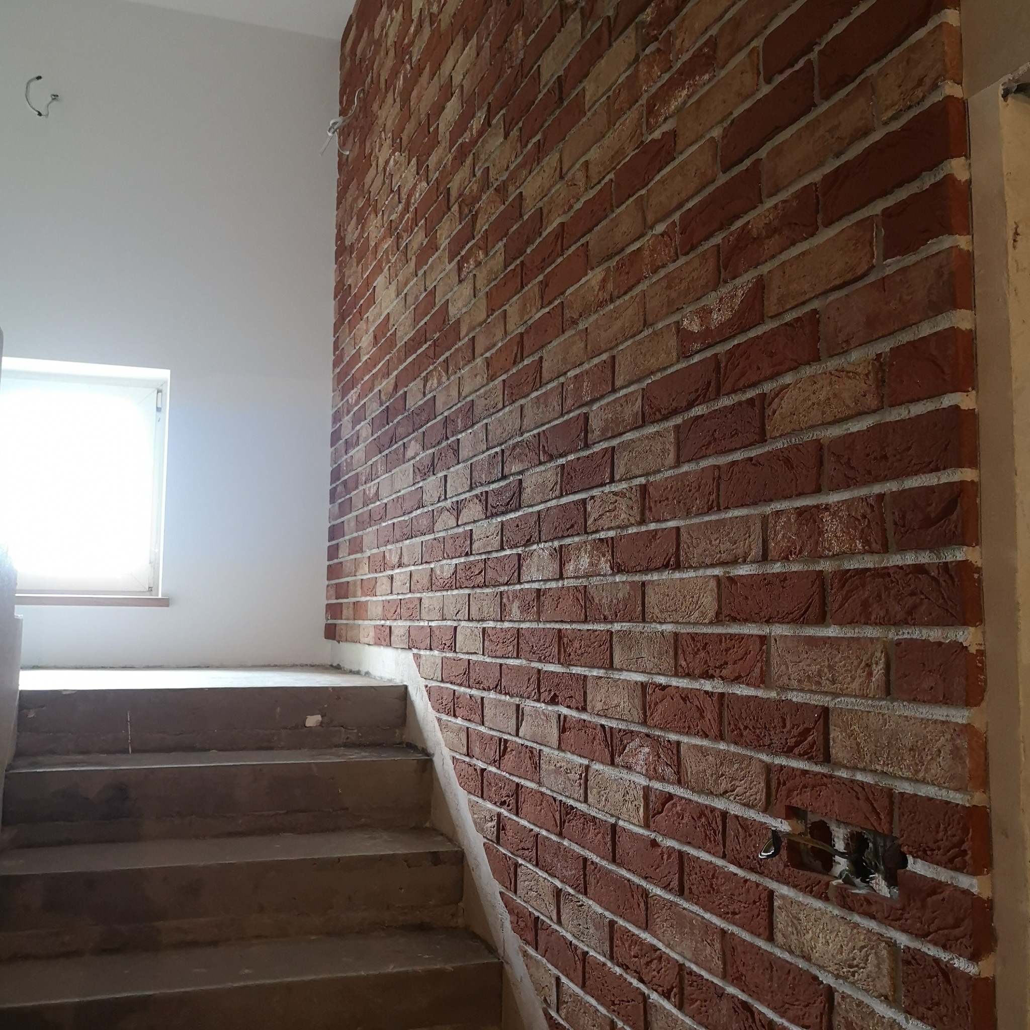 Ściana wykończona cegłą dekoracyjną w odcieniach czerwieni i beżu, przylegająca do betonowych schodów w jasnym pomieszczeniu z oknem, widoczne kable wystające ze ściany.