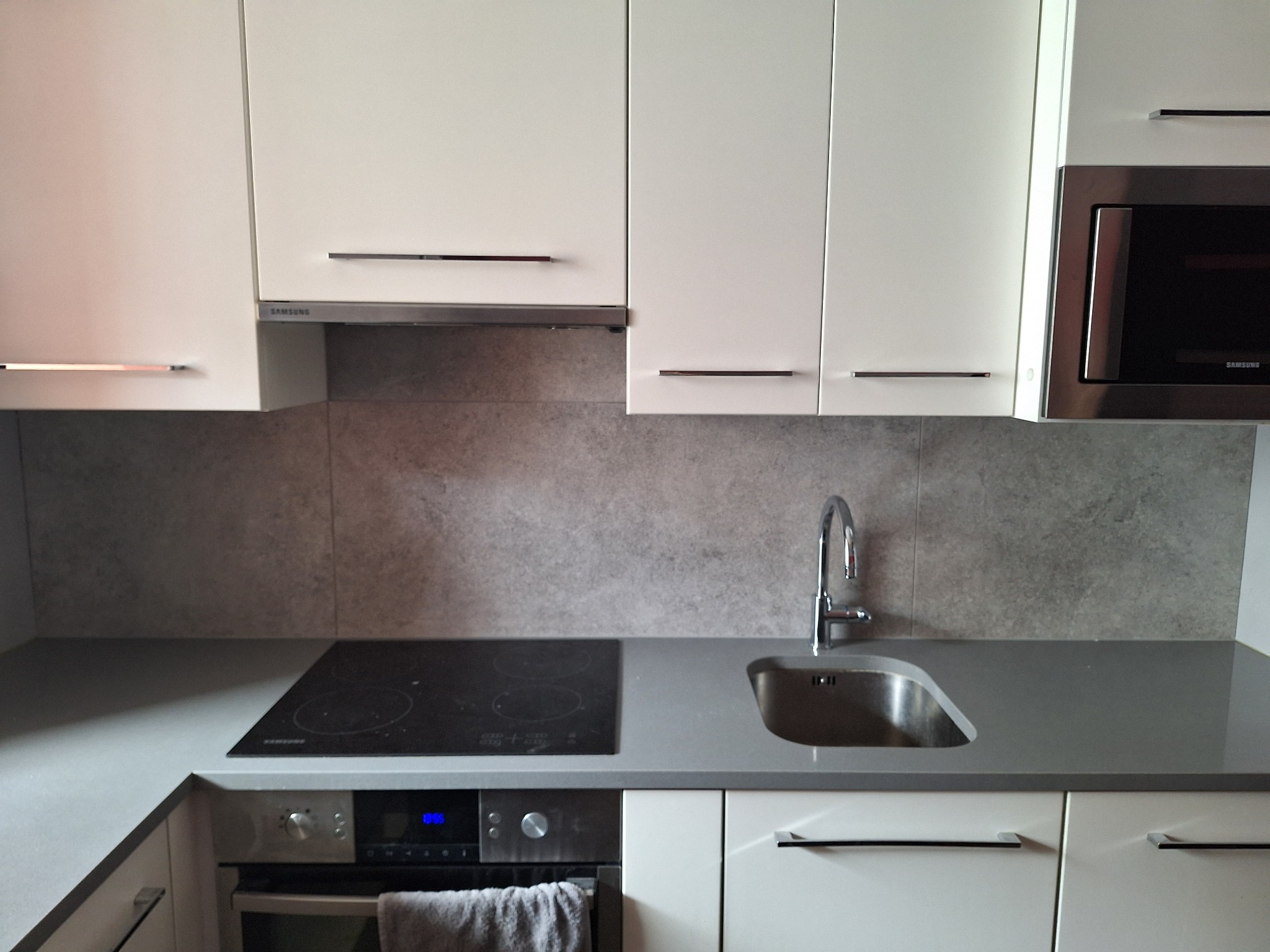 Nowoczesna kuchnia z białymi szafkami, szarym blatem, płytą indukcyjną i zlewozmywakiem ze stali nierdzewnej pod chromowaną baterią.