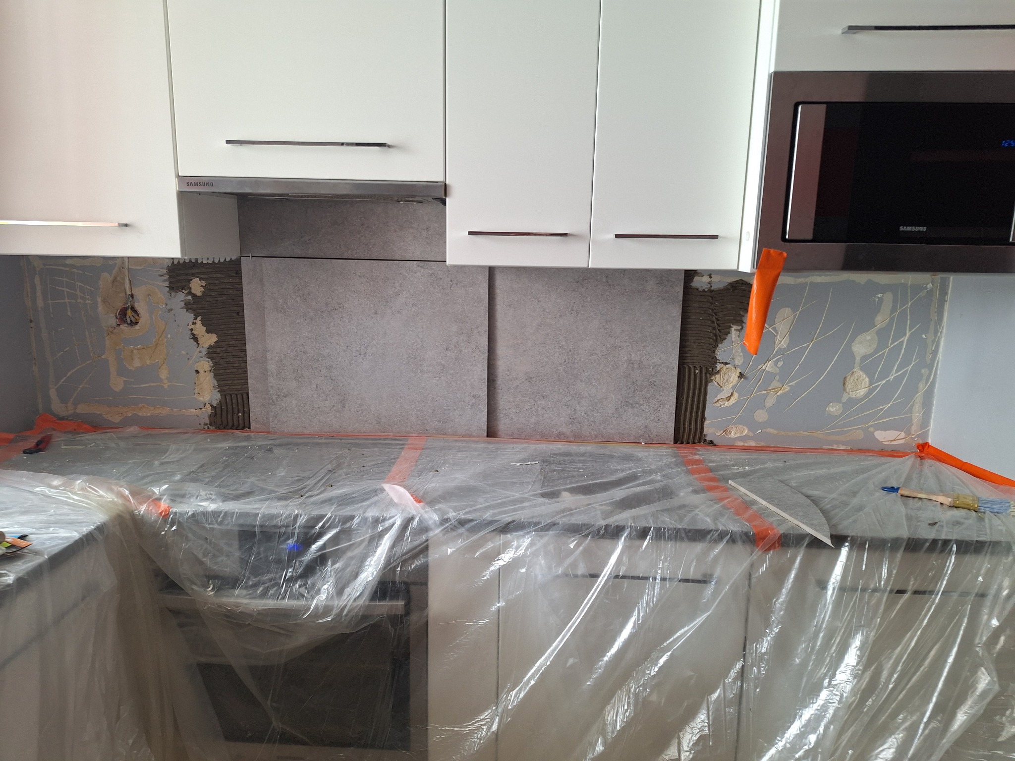 Montaż szarych płytek na ścianie nad blatem kuchennym, zabezpieczonym folią ochronną; widoczny fragment ściany z resztkami kleju i puszka elektryczna, białe szafki wiszące i mikrofalówka Samsung