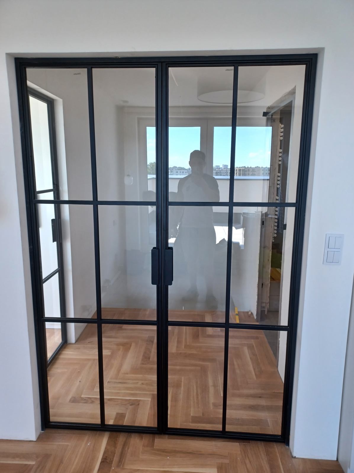 Nowoczesne dwuskrzydłowe drzwi szklane z czarnymi szprosami, widok na parkiet ułożony w jodełkę, odbicie sylwetki osoby w szybie.