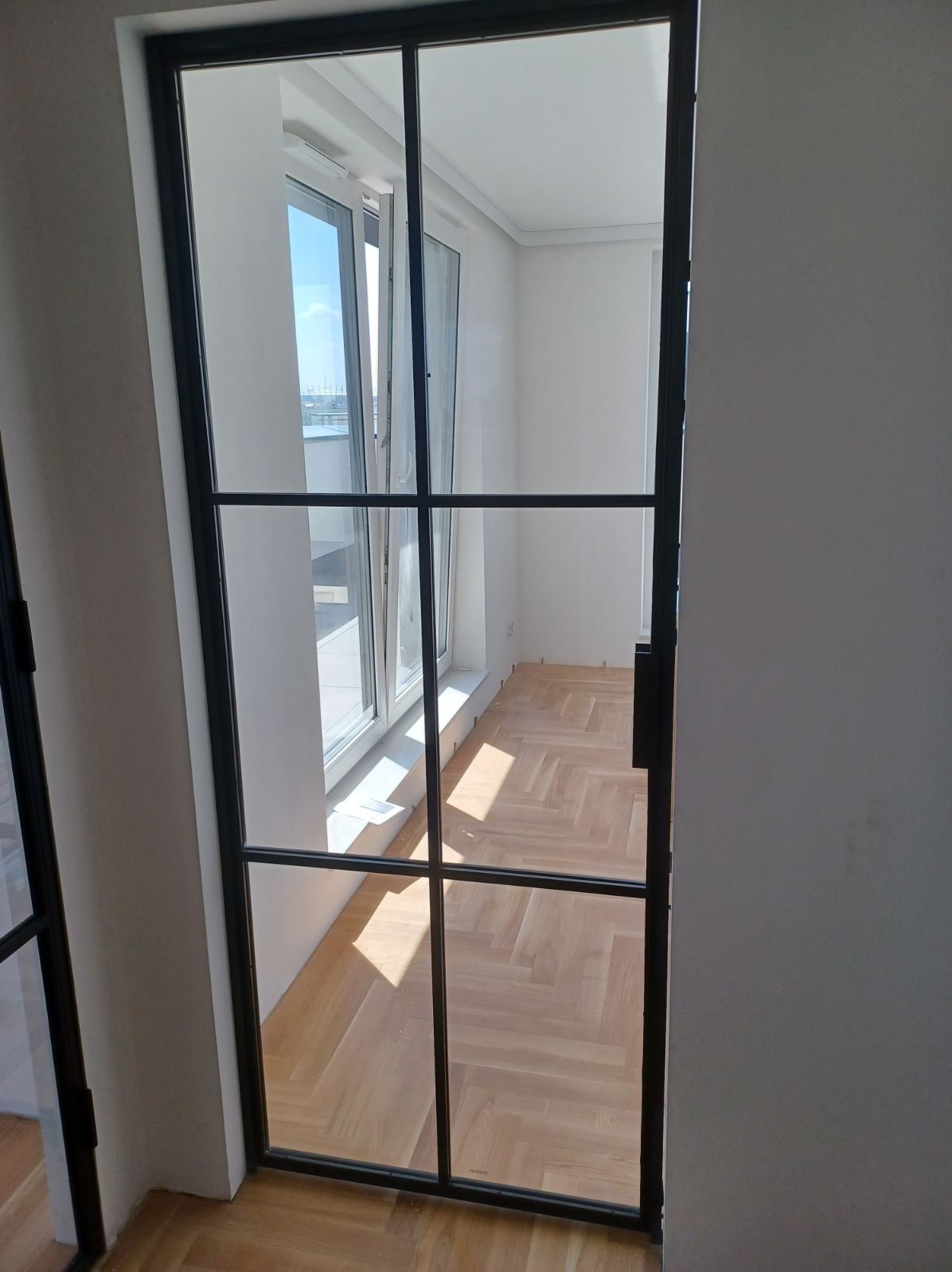 Nowoczesne, przeszklone drzwi wewnętrzne z czarną ramą, widok na parkiet ułożony w jodełkę i okno balkonowe z widokiem na miasto w oddali.