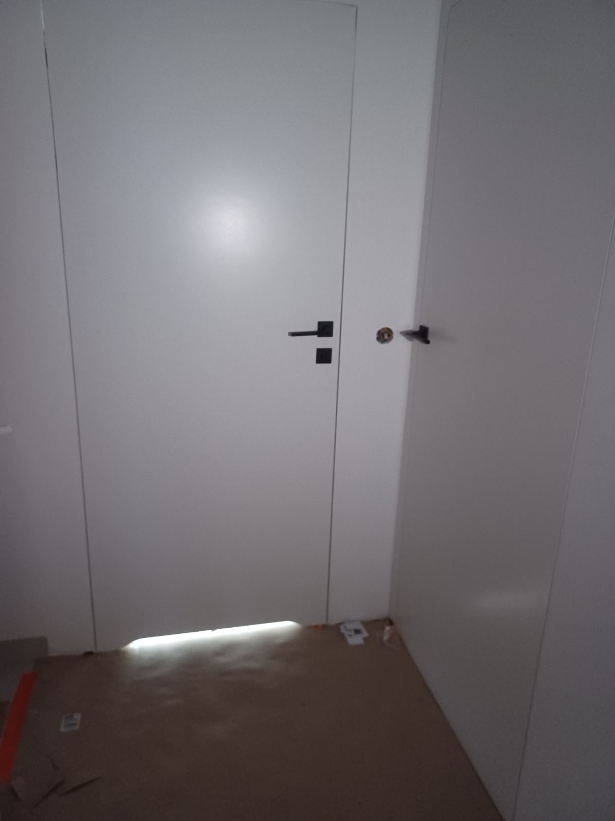 Białe drzwi wewnętrzne z czarną klamką i zamkiem, widoczne szczelina pod drzwiami z prześwitem światła, podłoga zabezpieczona kartonem.