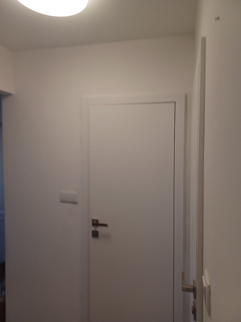 Białe, minimalistyczne drzwi wewnętrzne z prostą klamką i ościeżnicą, widoczne na tle białej ściany z włącznikiem światła.