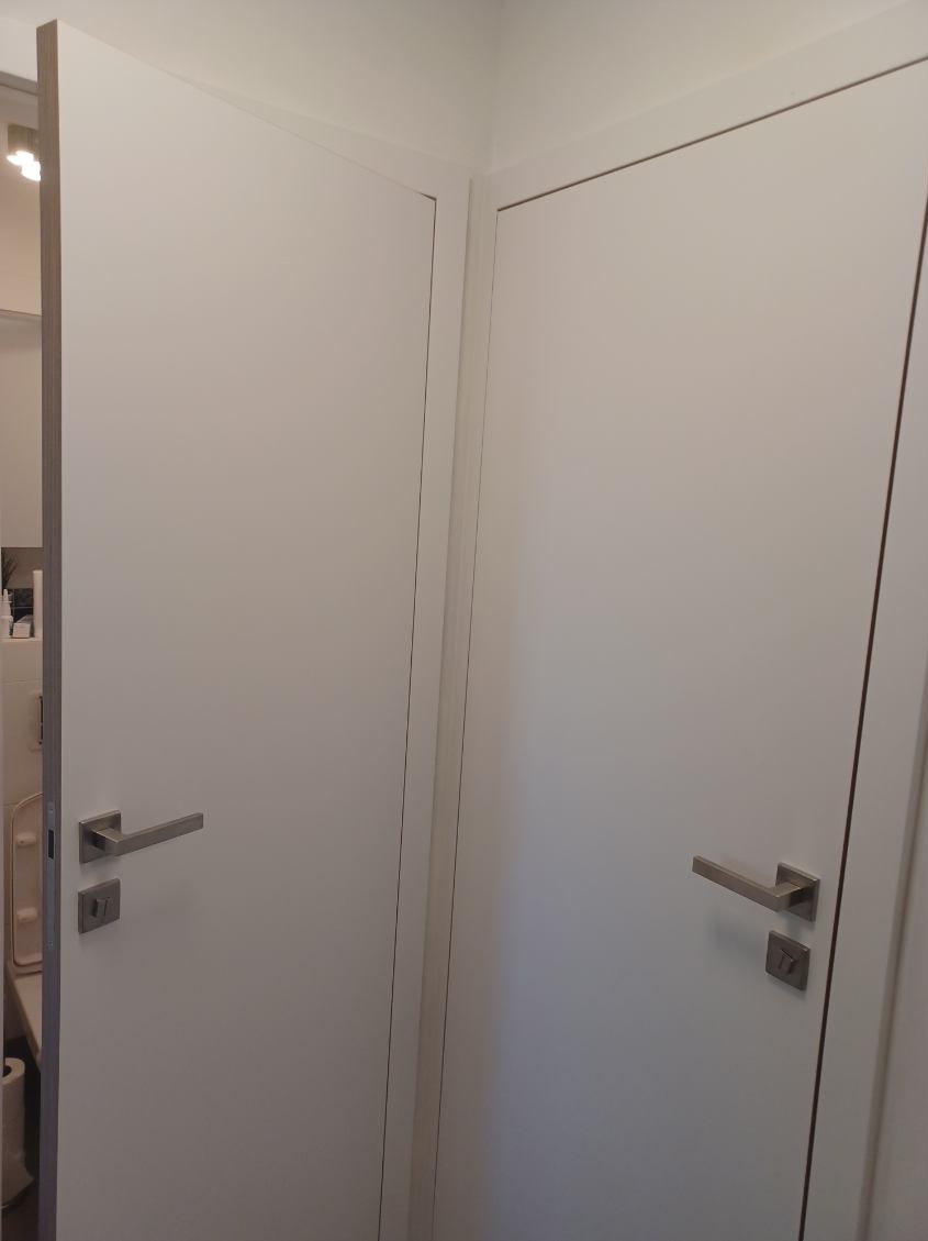 Dwa białe, nowoczesne drzwi wewnętrzne z minimalistycznymi, metalowymi klamkami, widoczne z bliska w rogu pomieszczenia, jeden z nich lekko uchylony, ukazujący fragment wnętrza.