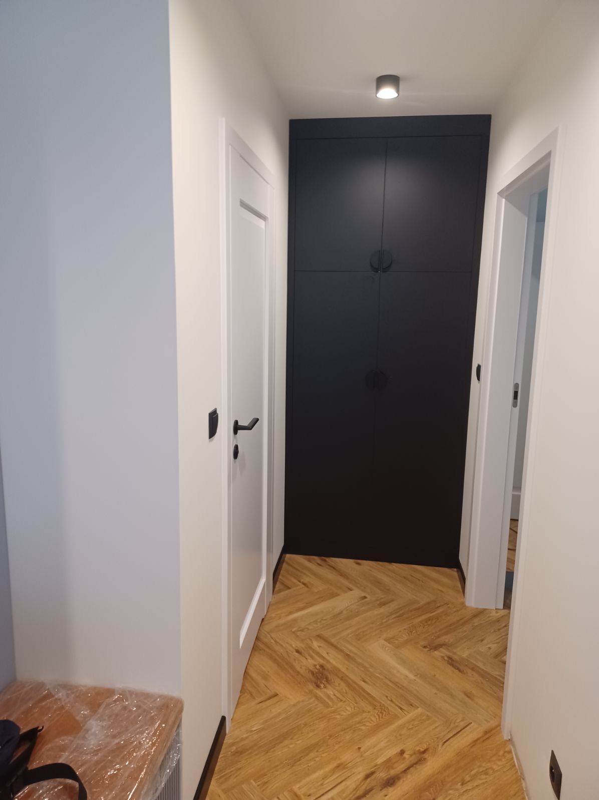 Wnętrze korytarza z jasną podłogą ułożoną w jodełkę, białe drzwi z czarnymi klamkami po lewej stronie, oraz duża, czarna szafa w zabudowie na wprost, oświetlona punktowym światłem.