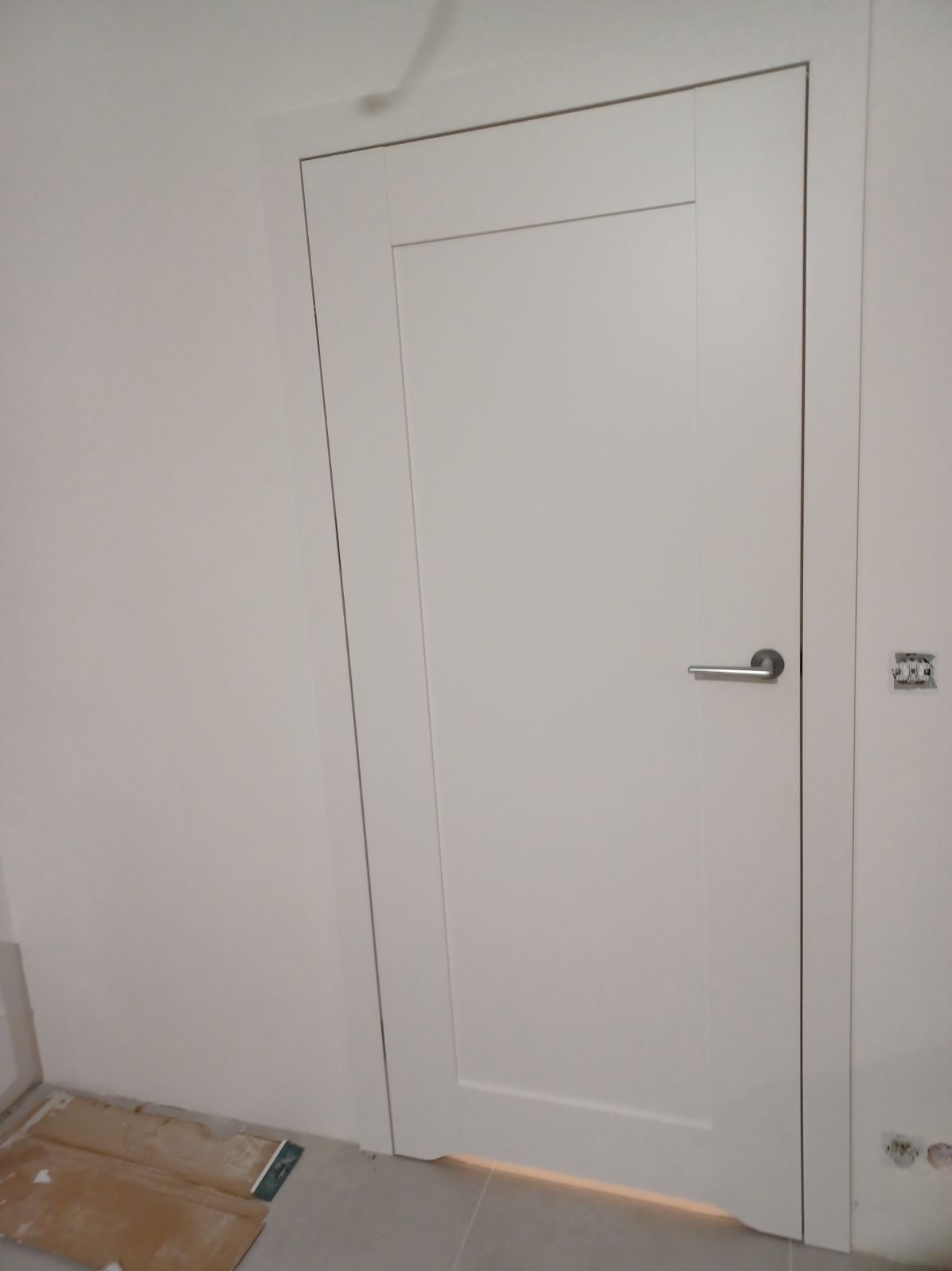 Zamknięte, białe drzwi wewnętrzne z minimalistyczną, srebrną klamką, osadzone w białej futrynie, z widocznym fragmentem podłogi i ściany z wystającą instalacją elektryczną.