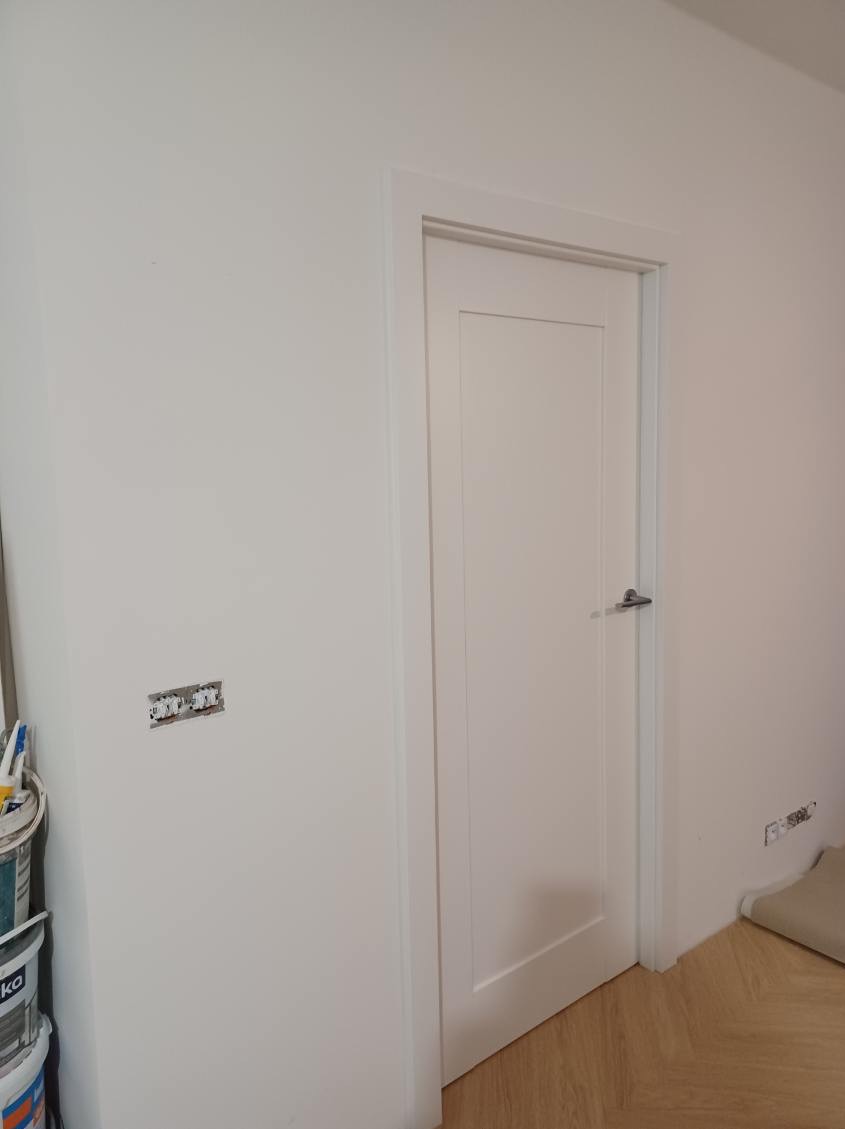Białe, minimalistyczne drzwi wewnętrzne z srebrną klamką, widoczne puszki elektryczne w ścianie i wiadro z farbą w rogu pomieszczenia.