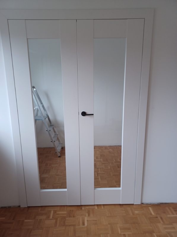 Białe, przeszklone drzwi wewnętrzne z czarną klamką, widoczna drabina w odbiciu szkła, parkiet na podłodze.