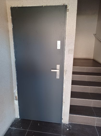 Szare, metalowe drzwi wejściowe w bloku, widoczne schody wyłożone czarno-szarą terakotą, ściana z widocznymi niedoskonałościami tynku.