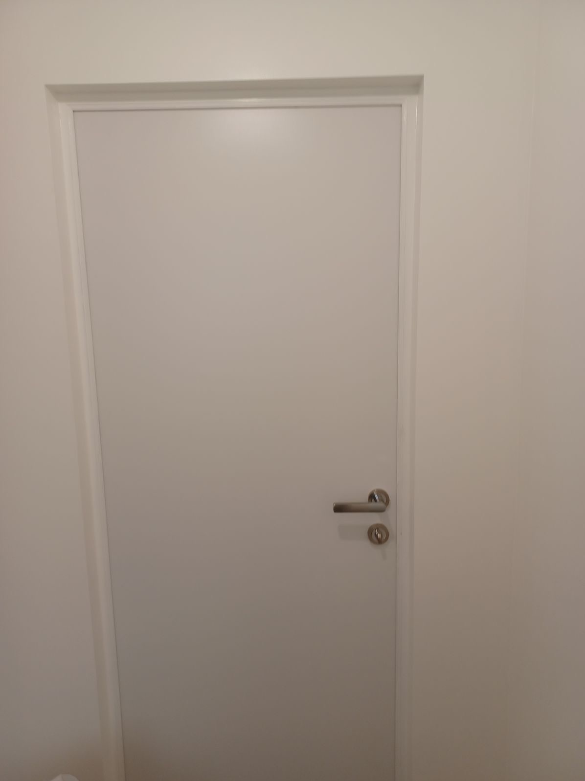 Białe drzwi wewnętrzne z srebrną klamką na tle białej ściany, widoczne ościeżnice.