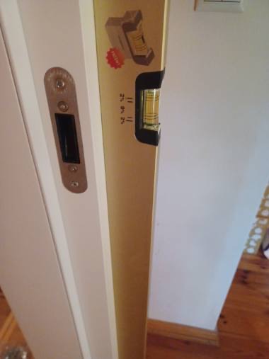 Poziomica przykładana do białych drzwi z zamkiem podczas montażu, widoczna drewniana podłoga.