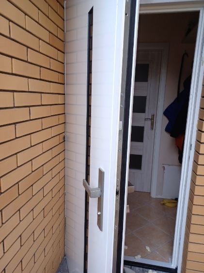 Otwarta biała, częściowo zamontowana, zewnętrzna drzwi wejściowe z metalową klamką, widoczne z zewnątrz budynku z cegły.