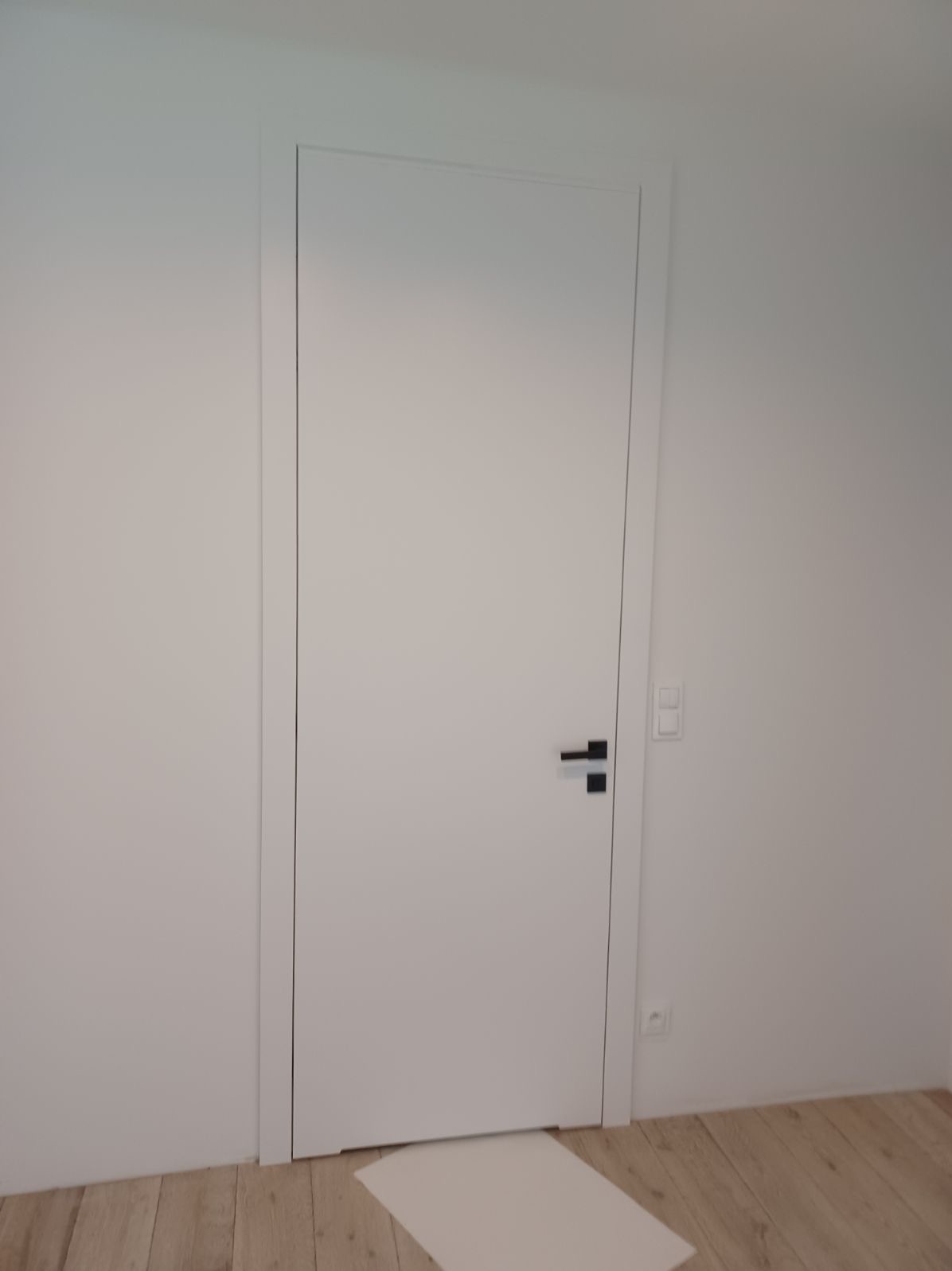 Białe, minimalistyczne drzwi wewnętrzne z czarną klamką na tle białej ściany i drewnianej podłogi. Pod drzwiami leży biała mata ochronna.
