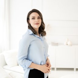 Services by Olesia Demianiuk - Portret kobiety w błękitnej koszuli i czarnych spodniach na tle jasnego, minimalistycznego wnętrza w Warszawie. Kobieta ma splecione dłonie i patrzy w obiektyw.