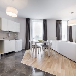 Services by Olesia Demianiuk - Jasne, przestronne wnętrze mieszkania z aneksem kuchennym, białymi meblami, stołem z krzesłami i dużym narożnikiem, oświetlone nowoczesnymi lampami wiszącymi.