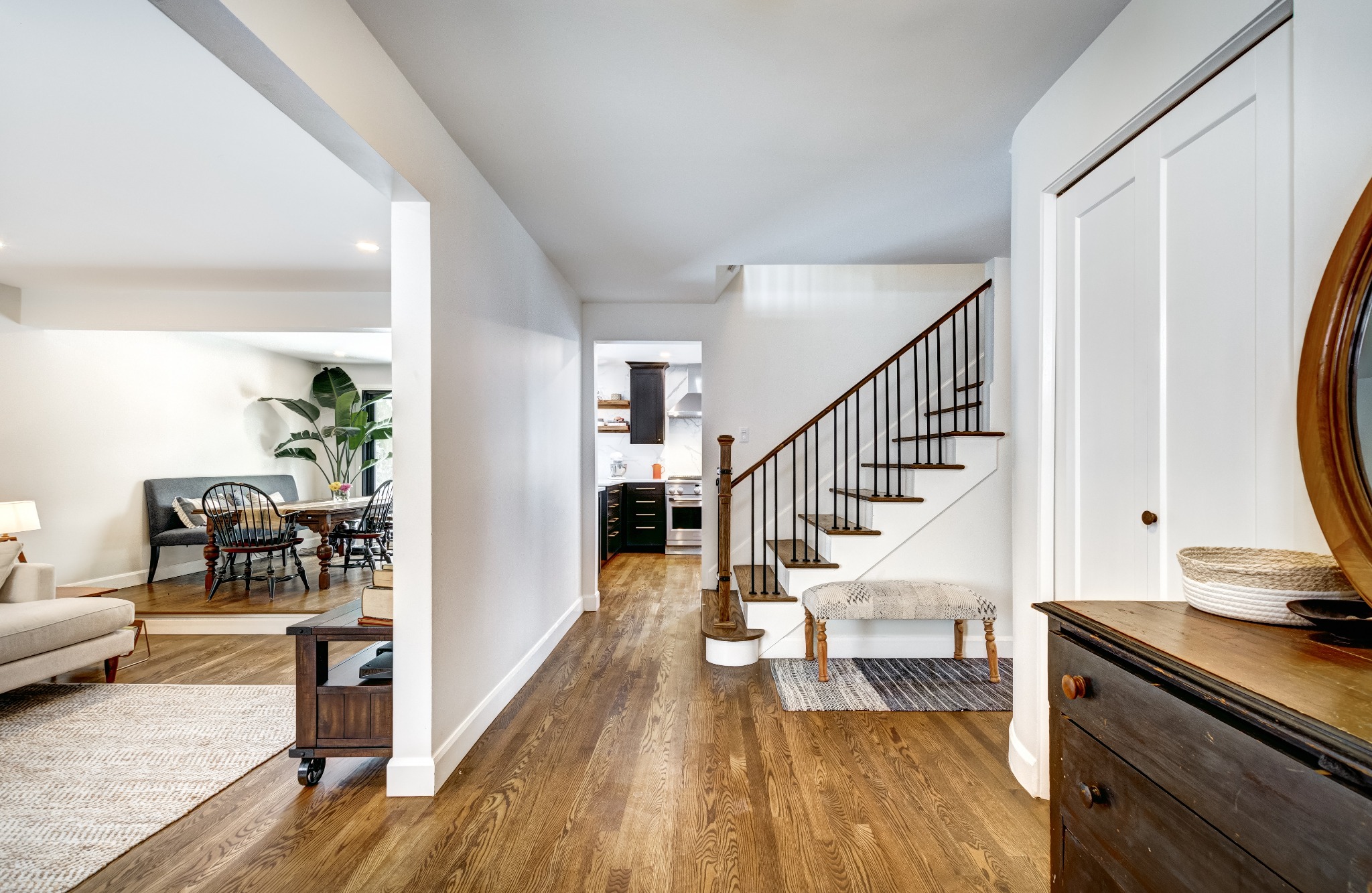 Jasne wnętrze domu z drewnianą podłogą, schodami z czarną balustradą, widokiem na salon i kuchnię, oraz antyczną komodą z okrągłym lustrem.