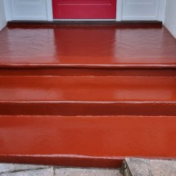 Filip Krzaczek - Pomarańczowo-czerwone schody wejściowe po świeżym malowaniu, widoczne detale ceglanej kolumny i płytki na górnym podeście ułożone w jodełkę.