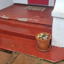 Filip Krzaczek - Czerwone schody wejściowe z odpryskującą farbą, doniczki z kamieniami po bokach. Zdjęcie 'Przed'.