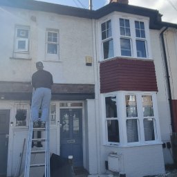 Filip Krzaczek - Pracownik na drabinie maluje elewację szeregowego domu z charakterystycznym oknem wykuszowym, widoczne numery domów 116 i 118.