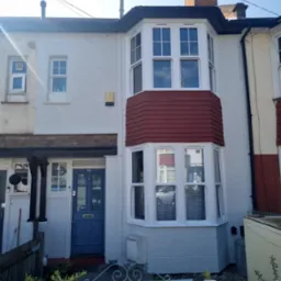 Odnowiona fasada szeregowego domu z niebieskimi drzwiami wejściowymi i białym oknem wykuszowym z czerwonymi płytkami, widoczne detale architektoniczne i świeże malowanie.