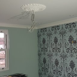 Filip Krzaczek - Pokój w trakcie remontu: ściana z tapetą w kwiaty, okno z widokiem na domy, wiszące kable elektryczne i ozdobny rozet na suficie.