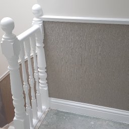 Filip Krzaczek - Fragment klatki schodowej po odświeżeniu: biała balustrada z ozdobnymi tralkami, ściana z tapetą w odcieniach szarości, białe listwy przypodłogowe i panel ścienny.