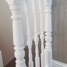 Filip Krzaczek - Biała, odnowiona balustrada schodowa z ozdobnymi tralkami i kulistymi zakończeniami, widok z góry na dywan na schodach i fragment ściany z tapetą.