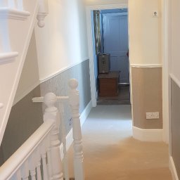 Filip Krzaczek - Odnowione wnętrze korytarza z pomalowanymi ścianami na biało i szaro, widoczne schody z białymi balustradami oraz ozdobna lampa wisząca.