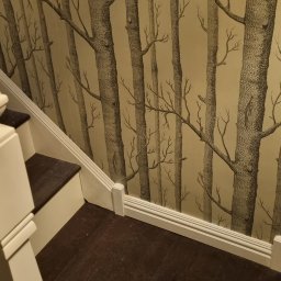 Filip Krzaczek - Klatka schodowa z tapetą w motywie drzew, drewniane schody z białymi balustradami i brązowymi stopniami.