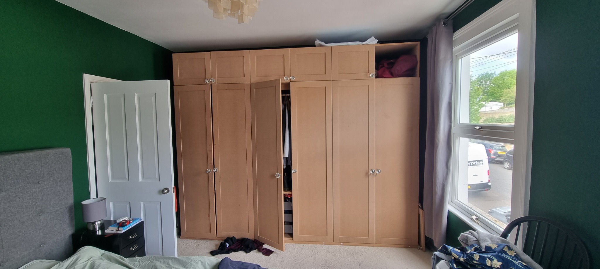 Pokój z ciemnozieloną ścianą, jasnymi panelami podłogowymi i dużą, beżową szafą z otwartymi drzwiami, widok na wnętrze szafy z ubraniami, okno z widokiem na ulicę.