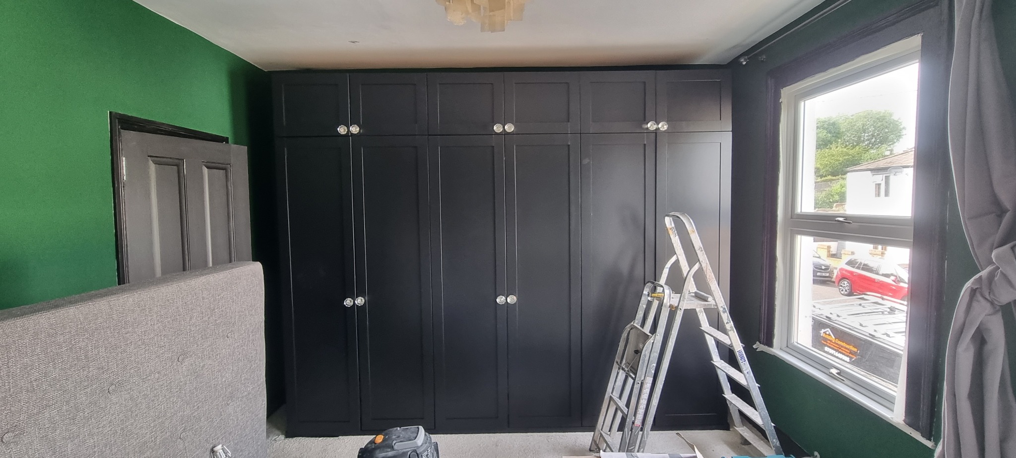 Wnętrze pokoju z ciemnozielonymi ścianami, zabudowaną szafą z czarnymi frontami i kryształowymi gałkami, częściowo zasłoniętą ramą łóżka i drabiną malarską przy oknie.