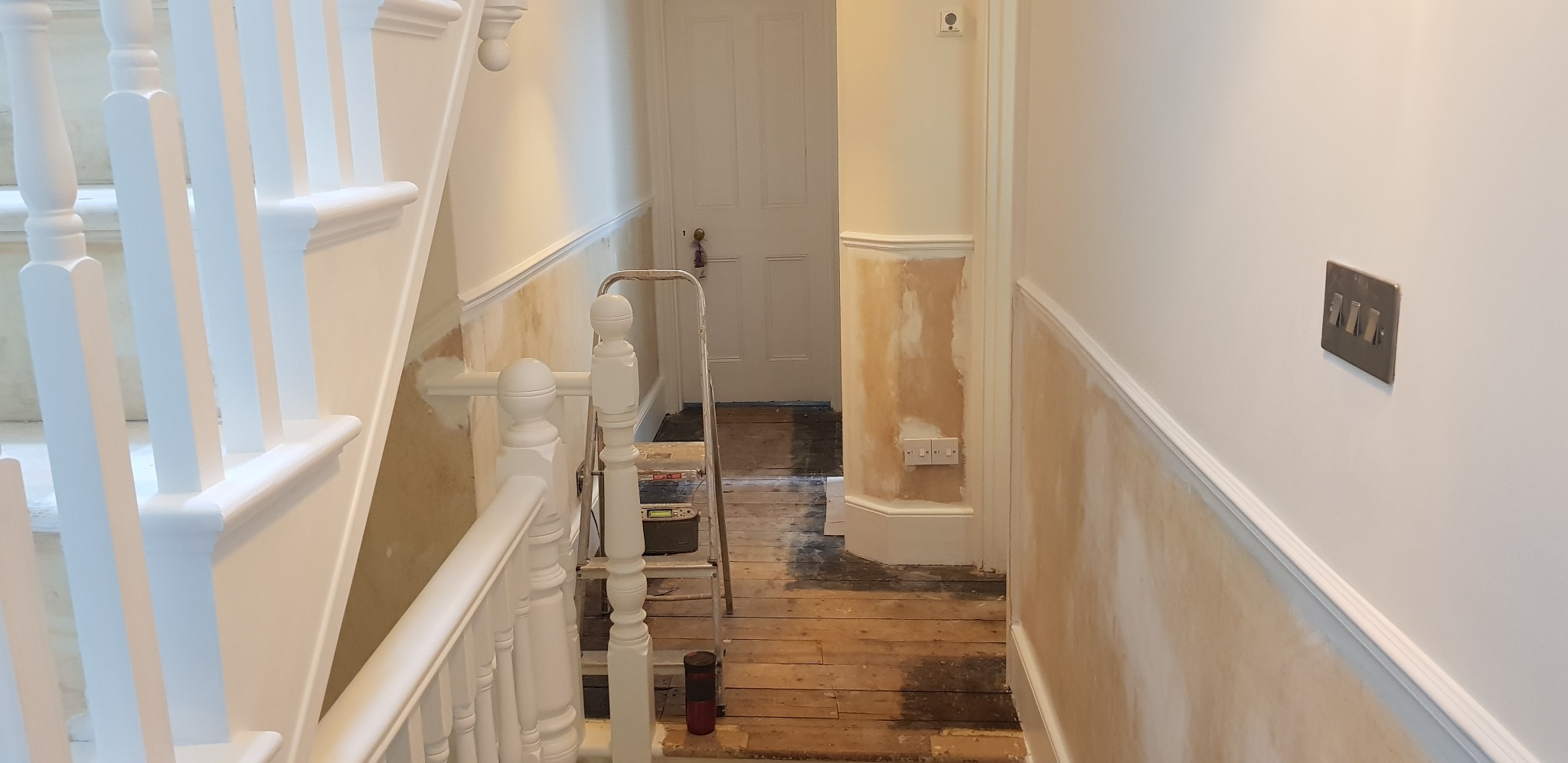 Korytarz w trakcie prac: ściany częściowo pokryte masą szpachlową, widoczna drabina, podłoga zabezpieczona folią, białe schody z balustradą.