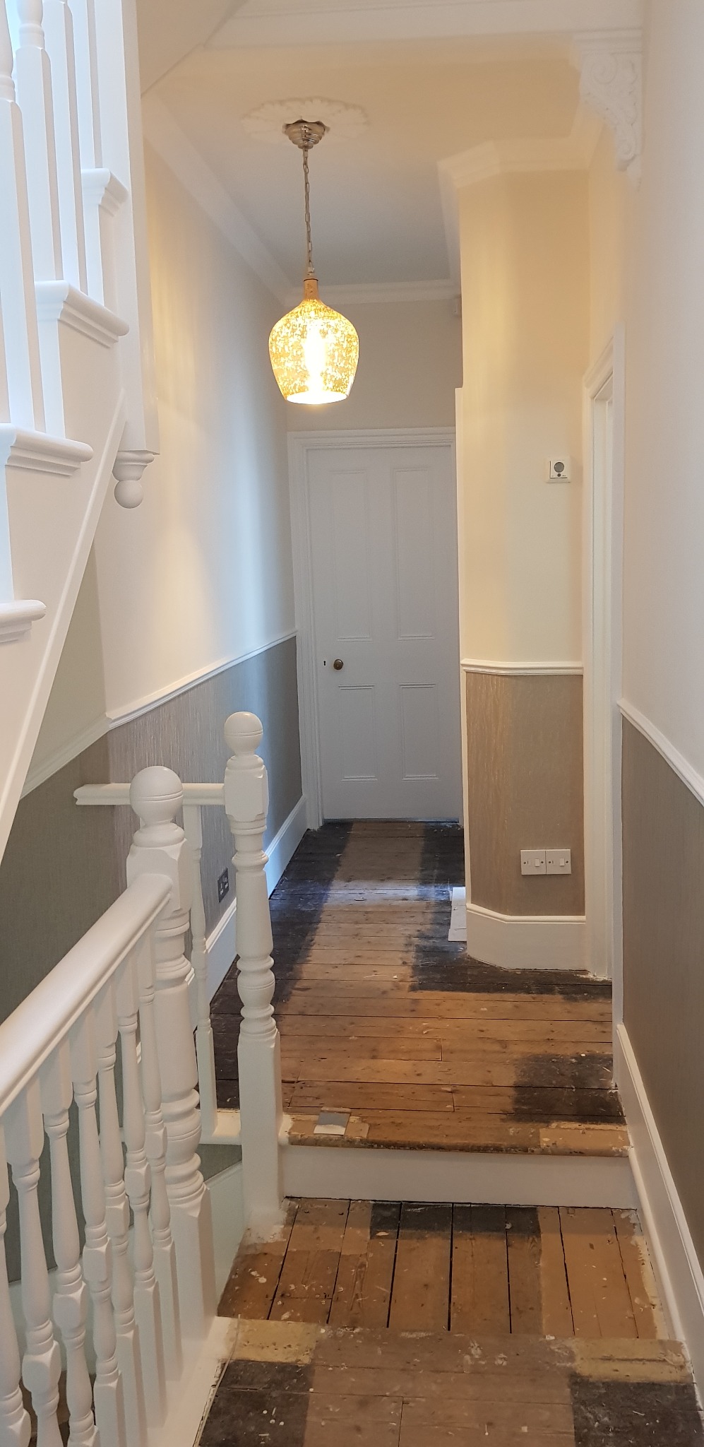 Korytarz w trakcie remontu: częściowo pomalowane ściany z boazerią, drewniana podłoga przygotowana do położenia paneli, widoczna lampa wisząca i biała balustrada schodów.