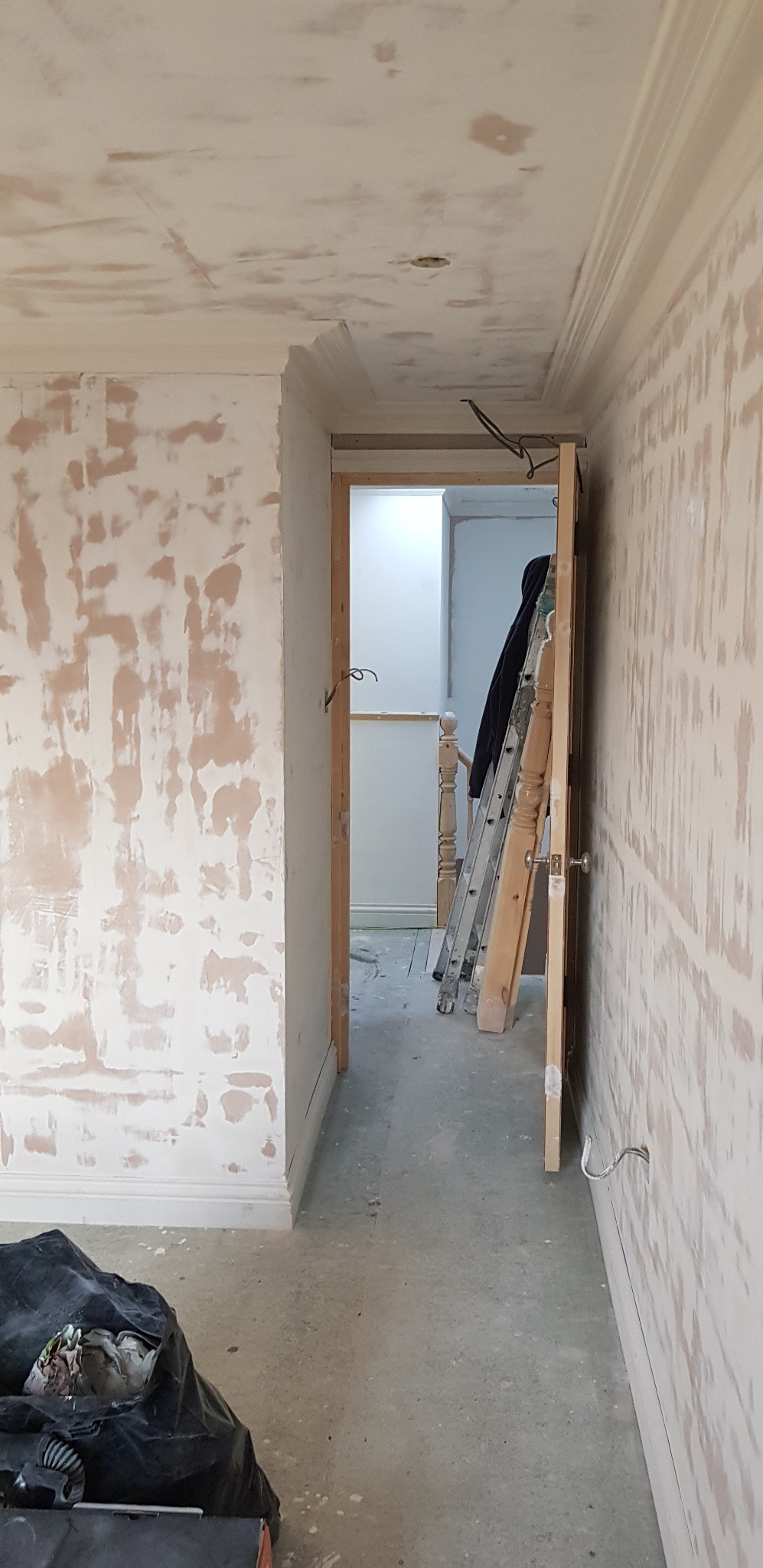 Wnętrze w trakcie prac przygotowawczych: ściany i sufit pokryte warstwą szpachli, widoczna drabina, otwarte drzwi do kolejnego pomieszczenia oraz worek z odpadami budowlanymi w rogu.