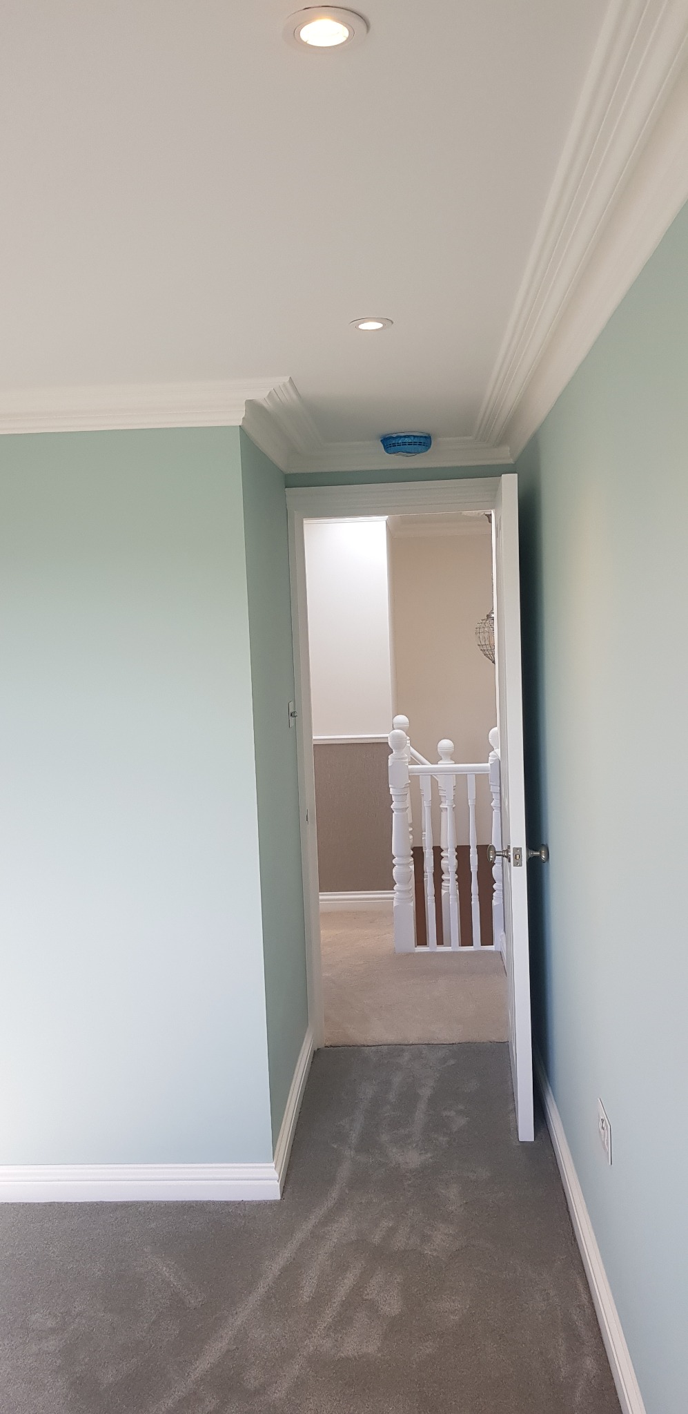 Świeżo wykończone wnętrze z pomalowanymi na pastelowy błękit ścianami, białym sufitem z zamontowanymi punktowymi lampami i białymi listwami przypodłogowymi. Widok na korytarz z szarą wykładziną...