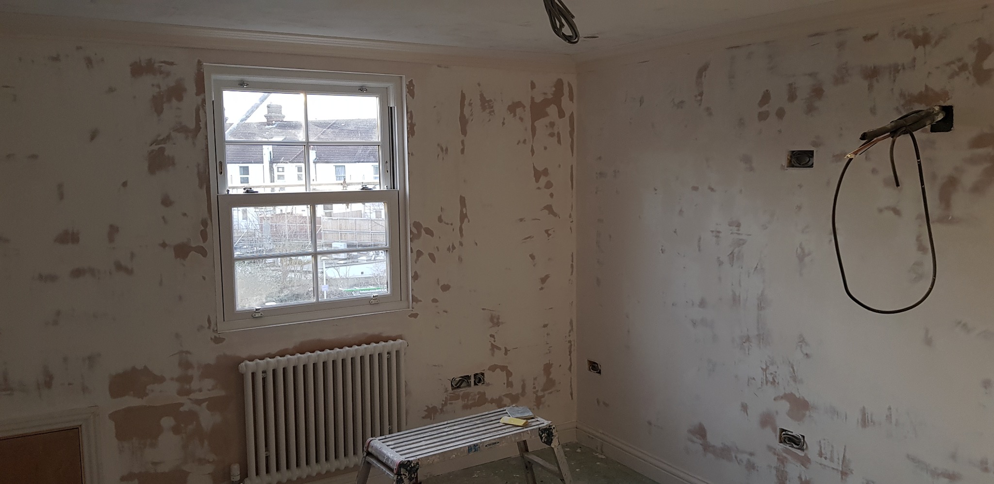 Pokój w trakcie przygotowywania ścian do malowania, widoczne ślady po szpachlowaniu, okno z widokiem na domy, grzejnik i drabina malarska z narzędziami.