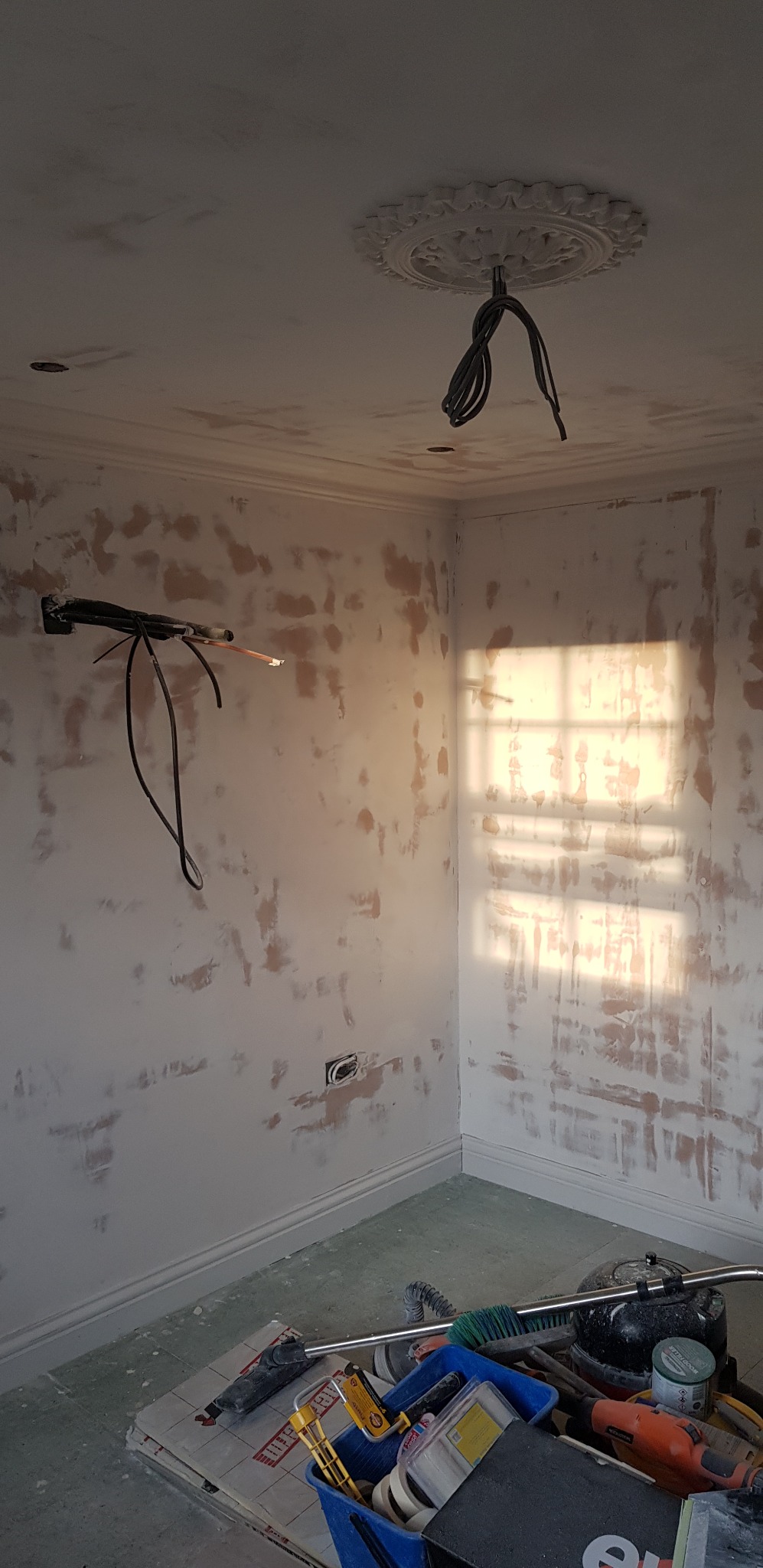 Pokój w trakcie prac przygotowawczych: ściany wstępnie wyszpachlowane, widoczne kable elektryczne wystające ze ściany i sufitu, rozproszone światło słoneczne przez okno, narzędzia malarskie...