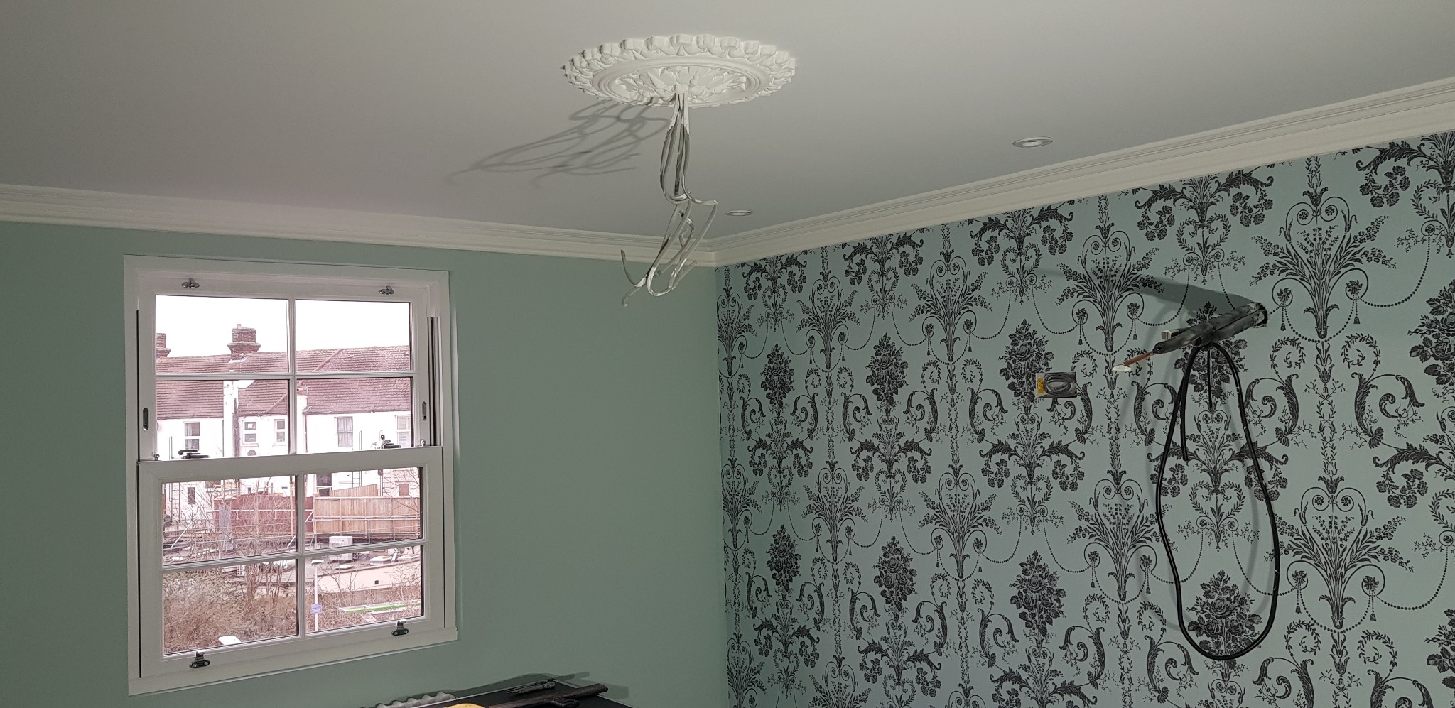 Pokój w trakcie remontu: ściana z tapetą w kwiaty, okno z widokiem na domy, wiszące kable elektryczne i ozdobny rozet na suficie.