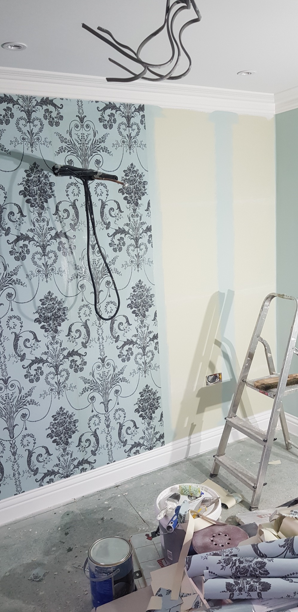 Ściana w trakcie odnawiania: połowa pokryta wzorzystą tapetą w odcieniach błękitu i szarości, druga połowa z fragmentami pomalowanymi na jasny, pastelowy kolor. Widoczne zwisające przewody...