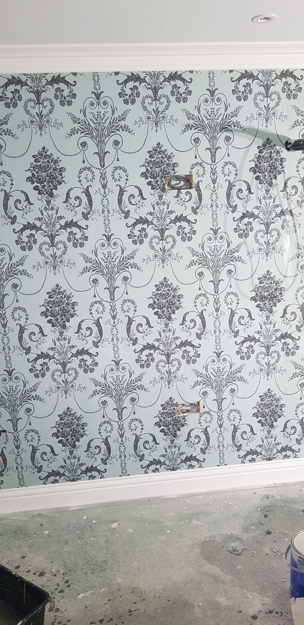 Ściana po położeniu jasnoniebieskiej tapety w ciemnoszary, kwiecisty wzór, widoczne puszki elektryczne, listwa przypodłogowa i fragment podłogi w trakcie remontu.