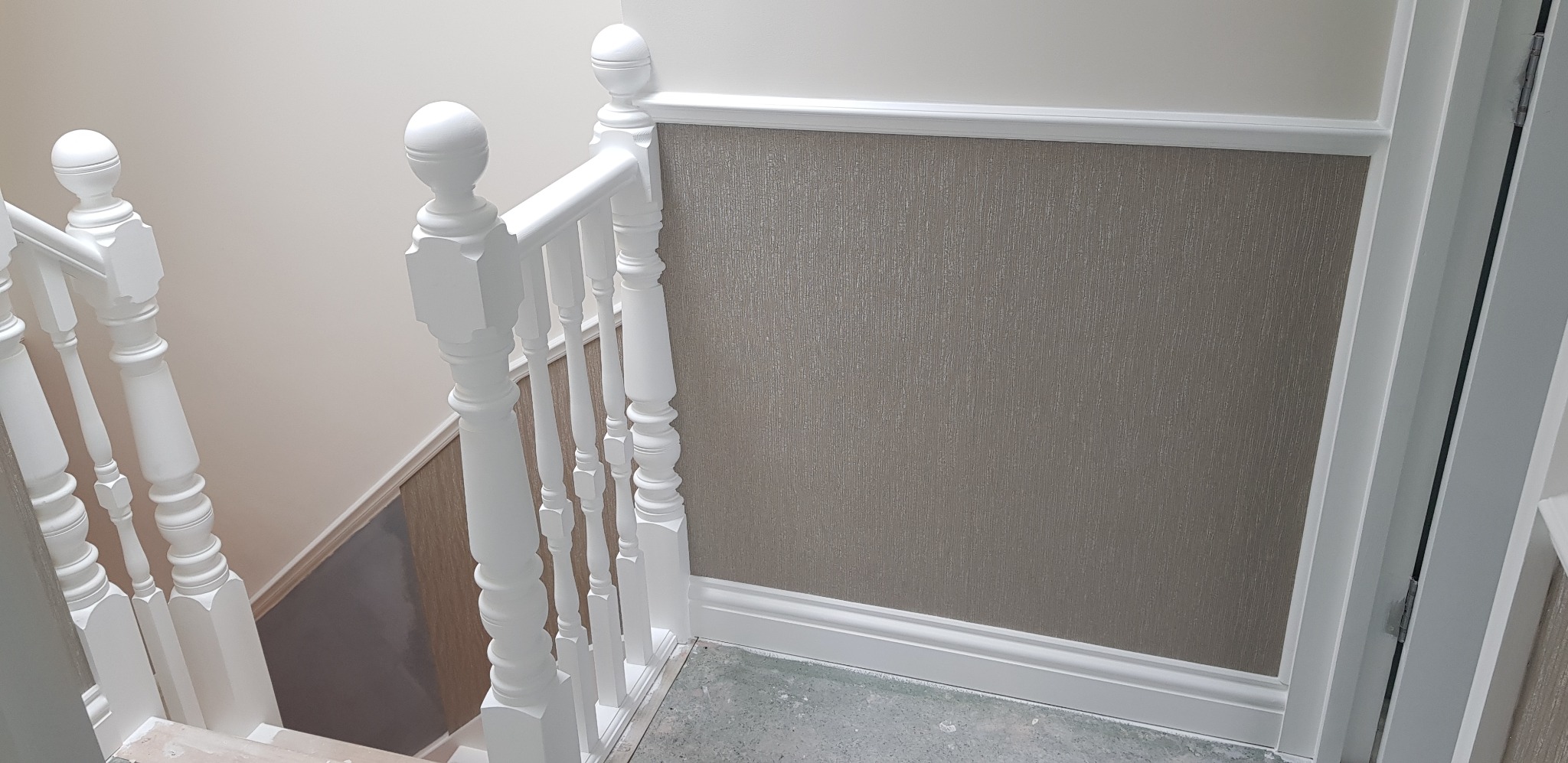 Fragment klatki schodowej po odświeżeniu: biała balustrada z ozdobnymi tralkami, ściana z tapetą w odcieniach szarości, białe listwy przypodłogowe i panel ścienny.