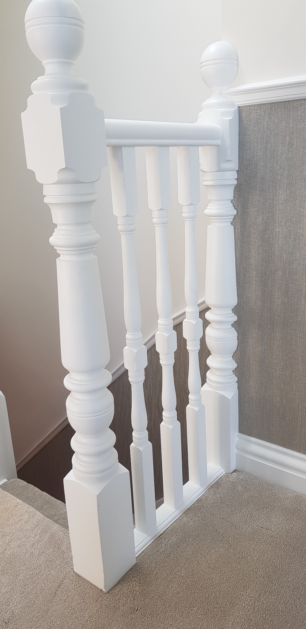 Biała, odnowiona balustrada schodowa z ozdobnymi tralkami i kulistymi zakończeniami, widok z góry na dywan na schodach i fragment ściany z tapetą.
